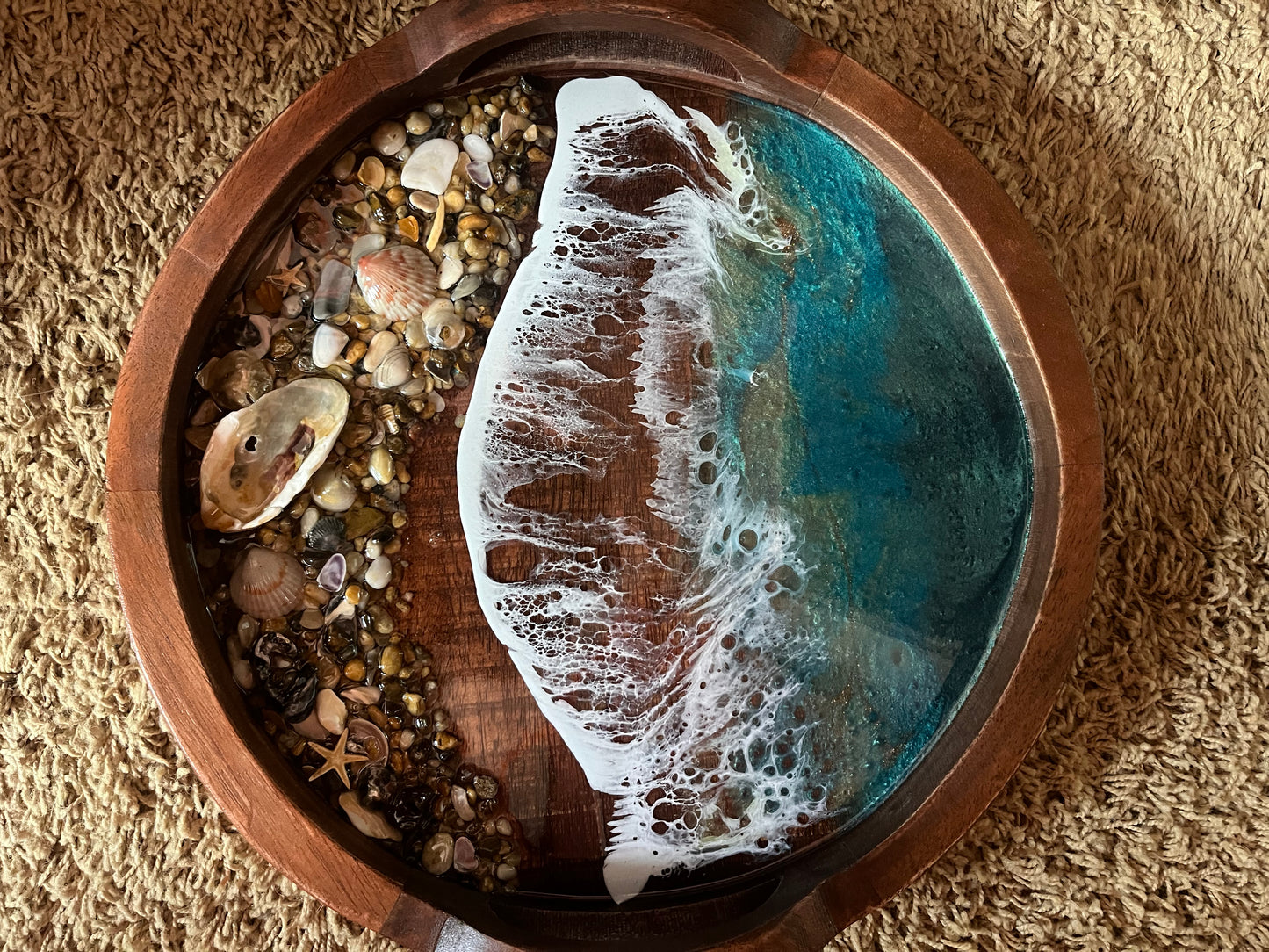 You need this gorgeous serving tray decorating your beach home. Beautifully hand crafted with resin waves memorializing the ocean crashing on the beaches of OBX.  Size is 14" Round Wood Tray with Handles.  Adorned with Ocean Resin Waves and Sea Shell Collected from the beaches of the Outer Banks. Serving Tray has Teal Blue Waves with Real Outer Banks Sea Shells. Hand made by Fyre Art Studio, an OBX Resin Artist located in Kill Devil Hills North Carolina.