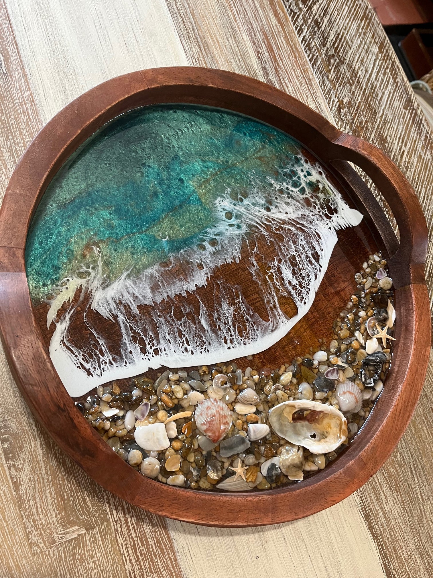 You need this gorgeous serving tray decorating your beach home. Beautifully hand crafted with resin waves memorializing the ocean crashing on the beaches of OBX.  Size is 14" Round Wood Tray with Handles.  Adorned with Ocean Resin Waves and Sea Shell Collected from the beaches of the Outer Banks. Serving Tray has Teal Blue Waves with Real Outer Banks Sea Shells. Hand made by Fyre Art Studio, an OBX Resin Artist located in Kill Devil Hills North Carolina.