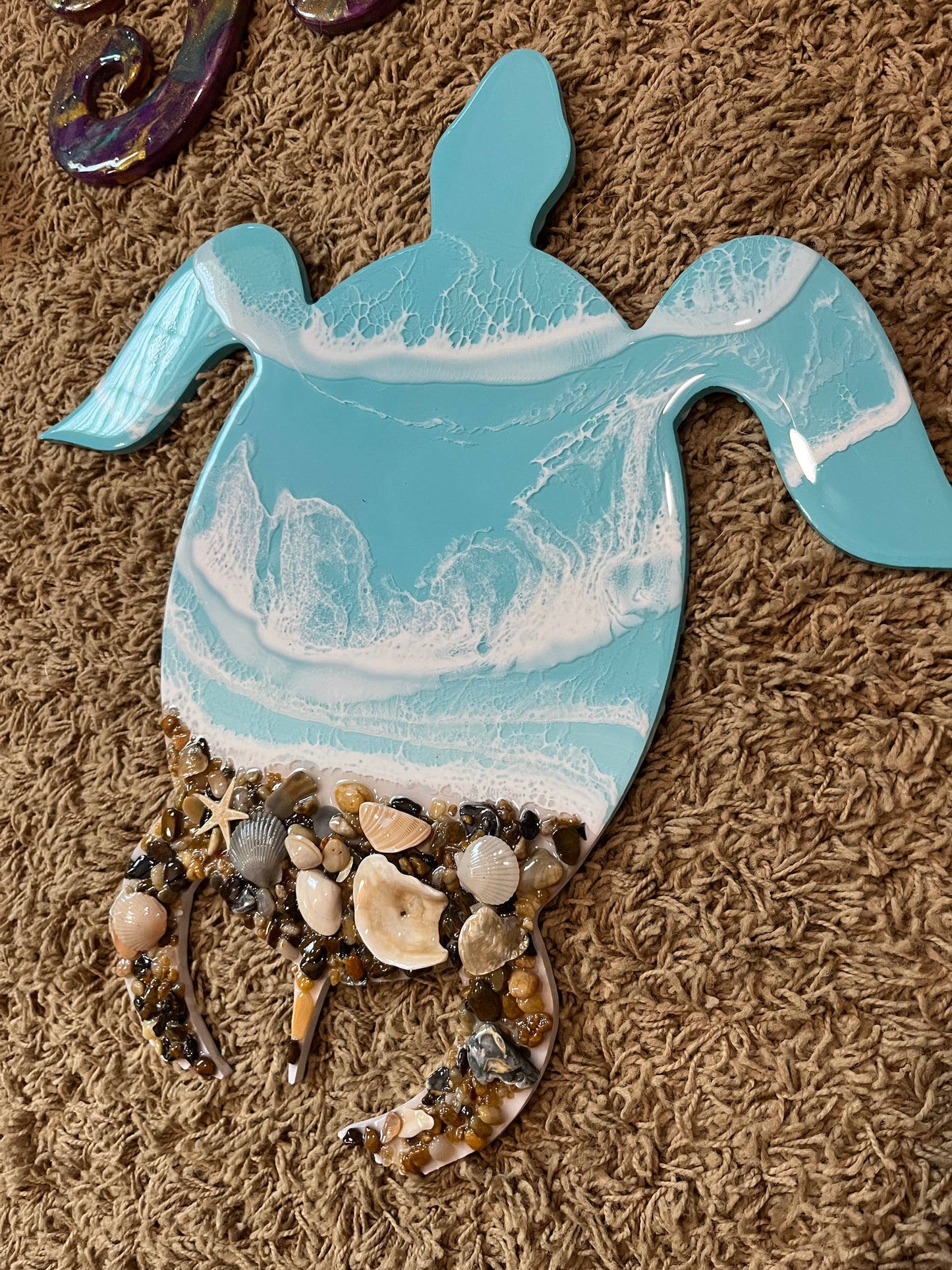 Light Blue Resin Waves Sea Turtle Wall Art with Real Outer Banks Shells on PVC sea turtle cut out. Real Outer Banks Shells. Size is 16" x 16" Take your love for the ocean to new, stylish depths with this unique Light Blue Resin Waves Sea Turtle Wall Art. Featuring real Outer Banks shells on a PVC sea turtle cut out, this piece adds a touch of quirky charm to any space. Measuring at 16" x 16", it's the perfect size to make a statement and bring a splash of the sea to your home.