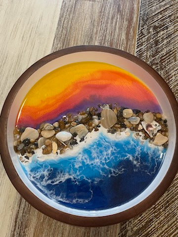 Size: 10" Round Wooden Serving Tray / Bowl  Dark wooden serving tray with white inside. Beautifully decorated with a colorful purple, orange and yellow resin sunset on the top half. Bottom half has deep blue resin ocean waves crashing onto real sea shells and polished stones collected from the beaches of the Outer Banks North Carolina. Fyre Art Studio Outer Banks Resin Artist.