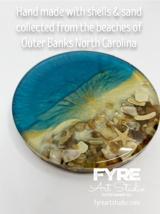 Original Shore Break Coaster with real OBX sea shells and beach rocks sealed in resin. Qty 1 Coaster - 4" diameter circle shape with cork backer.  Inspiration for the design comes from the beaches of the Outer Banks in North Carolina. Colored with shimmering mica powder in shades of medium blue, aqua teal 